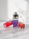 Three cosmetic lip gloss bottles from Lumiere Lip Oil arranged on a white marble surface against a white wall with soft leaf shadows. The center bottle is upright and filled with purple gloss, while the two side bottles lie on their sides in red and orange shades.